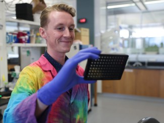 A blonde man in a the-dyed lab coat holds a square tray in one blue gloved hand