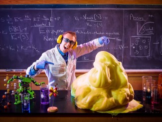 A man in a sequin-covered lab coat and blue rubber gloves stands in front of a chalk board covered with equations gestures at a large pile of yellow slime on a lab bench covered with beakers and models of molecules