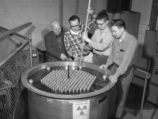 Four men stand over a cylindrical tank labeled with radiation warning. An older man on the left points towards the surface of water in the tank as two younger men hold a rod extending below the surface.