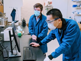 Man in blue lab coat wearing a mask looks at computer monitor as another man looks on
