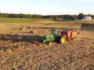 A green tractor pulls a red baler in a field of brown grass. Rectangular bales of grass are stacked in the background and scattered through the field. A line of grass and trees with a white barn are visible in the background. 