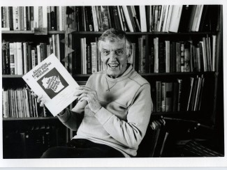 black and white photo of smiling man seated in front of a bookshelf holding a bound paper titled "Alternative energy futures for Wisconsin"