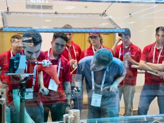 A man in a blue shirt leans in to observe as man in red shirt adjusts mechanism on a model wind turbine while a group of other men in red shirts look on