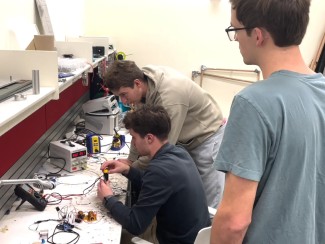 Three students work together at an electronics workbench, with one soldering a circuit while the others observe, surrounded by a power supply, soldering station, multimeter, and various wires and components.