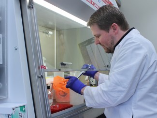 A man in a white lab coat and blue latex gloves pipettes liquid under a ventilation hood.