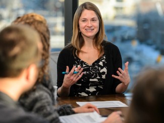 a woman smiles and gestures while speaking at a table with colleagues, whose backs are visible in the foreground, papers spread on the table in front of them, with a large window in the background.