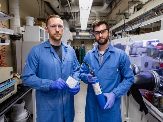 Two men in blue lab coats, gloves, and safety glasses pose holding objects in a laboratory