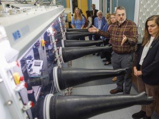 A man in a plaid shirt and safety glasses gestures toward a long glovebox with black gloves extending into the room, explaining it to a woman in a dark blazer, as a group of visitors looks on behind them.