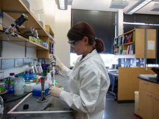 A woman in a lab coat and safety glasses uses a pipette at a lab bench cluttered with jars, tubes, and supplies, with bookshelves visible behind her.