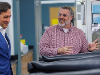 A man in a blue blazer looks on with a smile as a bearded man in a red checked shirt and safety glasses gestures animatedly beside a glovebox with black gloves in a laboratory.