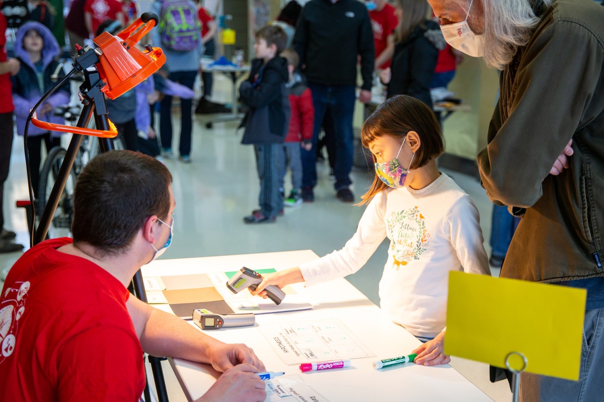 A girl uses a handheld laser thermometer to measure the temperature of various colored sheets on a table.