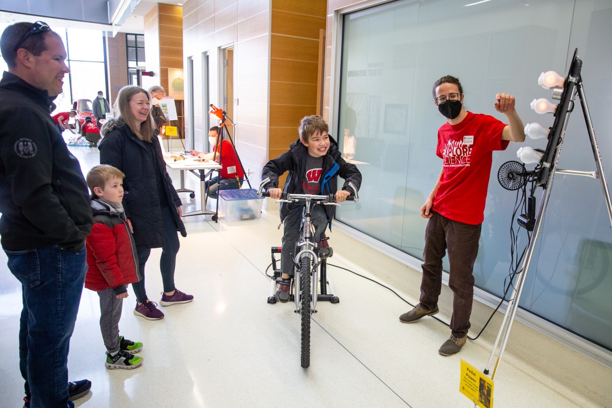 A kid smiles while powering lightbulbs by peddling a stationary bike while his family looks on.