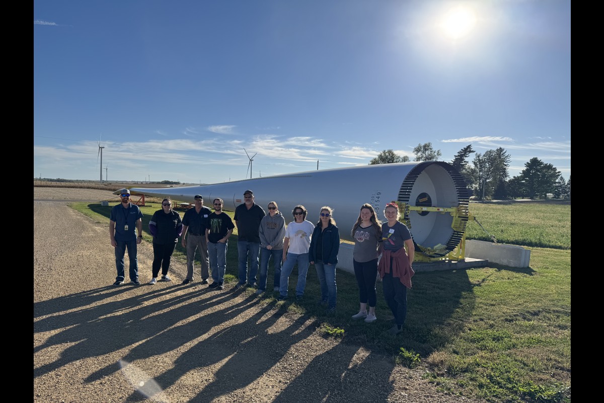 Teachers tour an operations and maintenance facility for a Wisconsin wind farm and stand next to a spare blade.