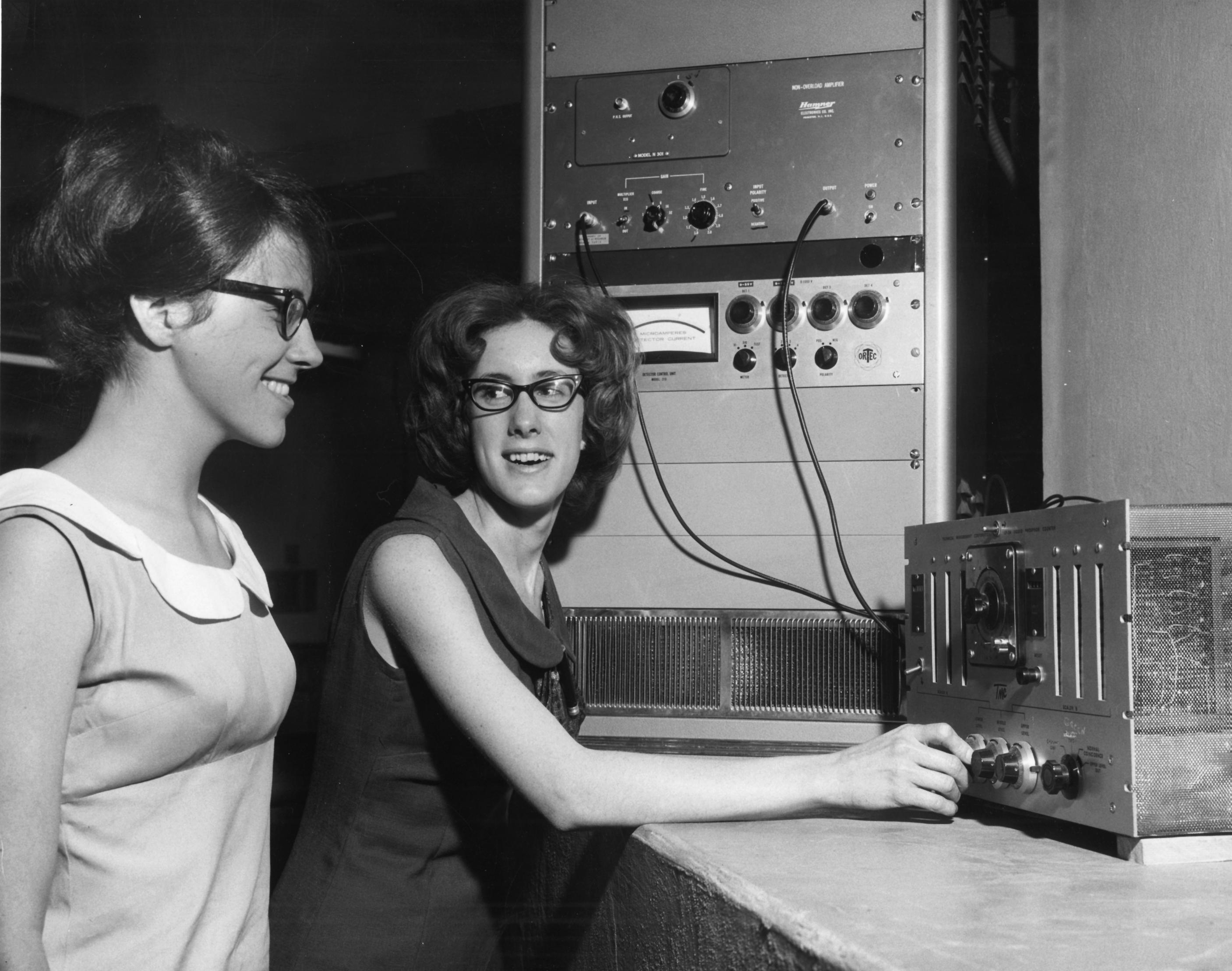 Black and white photo of two women. One is seated in front of a control panel and turned towards the other, who is observing over her shoulder